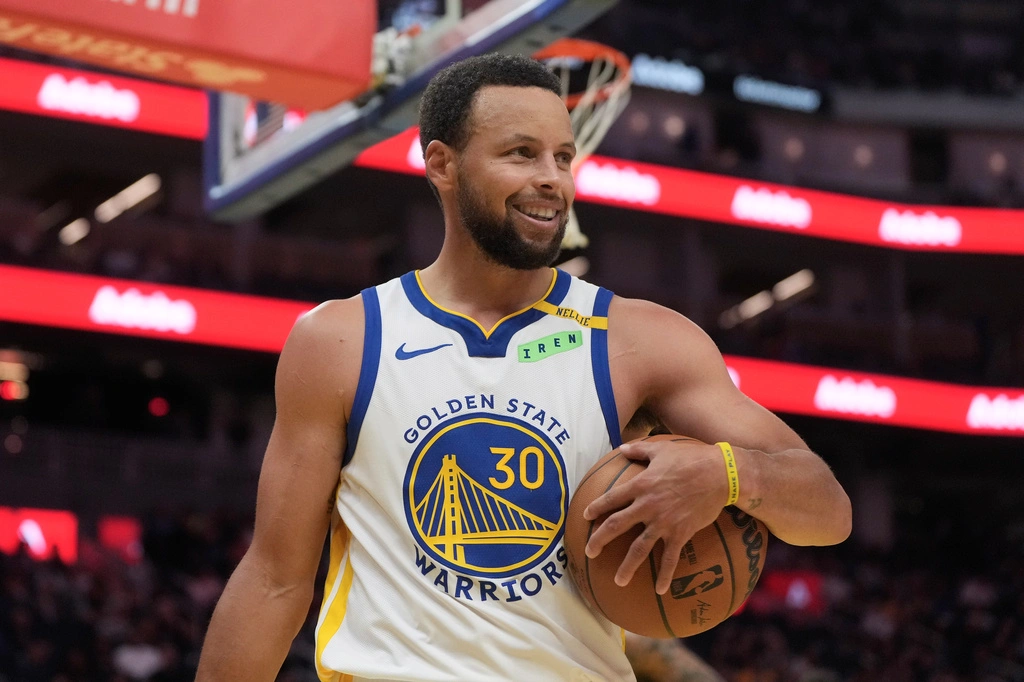 Stephen Curry in a white Golden State Warriors jersey smiles while holding a basketball during a preseason game against the Trail Blazers